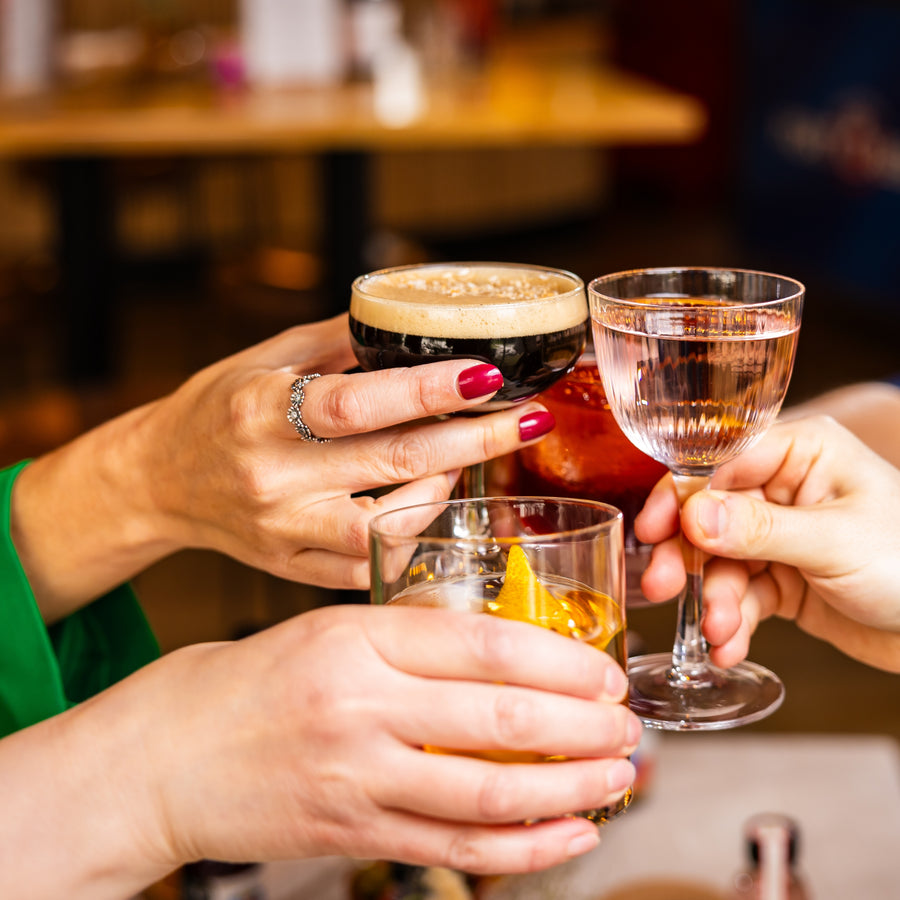 Three people are toasting in what looks like a bar. The picture is warm in colour. One lady holding the espresso martini has a bright green jacket on and red nails. There is an old fashioned and a lychee martini in shot.