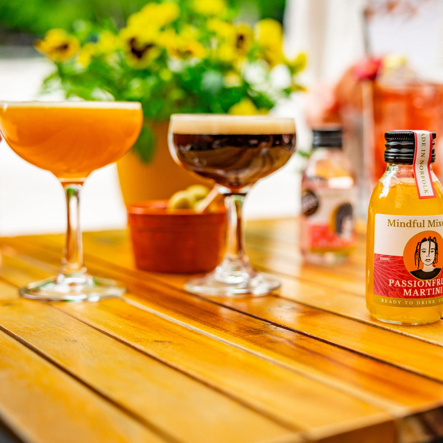 There is a passion fruit martini and espresso martini on an outdoor table, with a single-serve bottle of passionfruit martini. A blurred plant is in the background along with a bowl of olives. It looks like it is sunny outside.