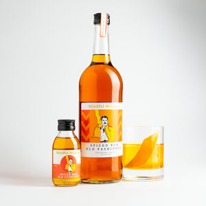 The small Spiced Rum Old Fashioned bottle is in the picture, alongside a large 750ml bottle. It is the same shape and similar label, but the bottle is a lot larger. Next to it is a glass of Spiced Rum Old Fashioned in a short tumbler. The cocktail is a caramel colour and has a diagonal strip of orange in the glass, and a large cube of ice. The background is white.