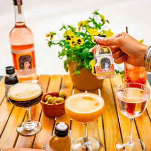 There are some cocktails on a table which looks like it is outside. A bright plant is on the table in a yellow plant pot with yellow flowers. A lady is holding the mini lychee martini bottle. An espresso martini, passionfruit martini and lychee martini are all on the table, with a bowl of olives and a large bottle of Lychee Martini.