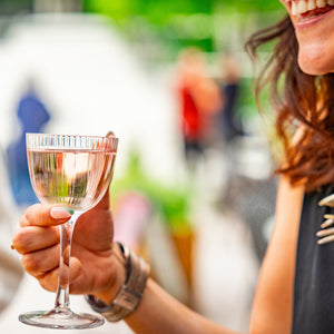 There is a lady holding a glass of Lychee Martini. She is smiling but the rest of her face is out of view. It looks like she is sitting outside, and the shot is bright and sunny. 