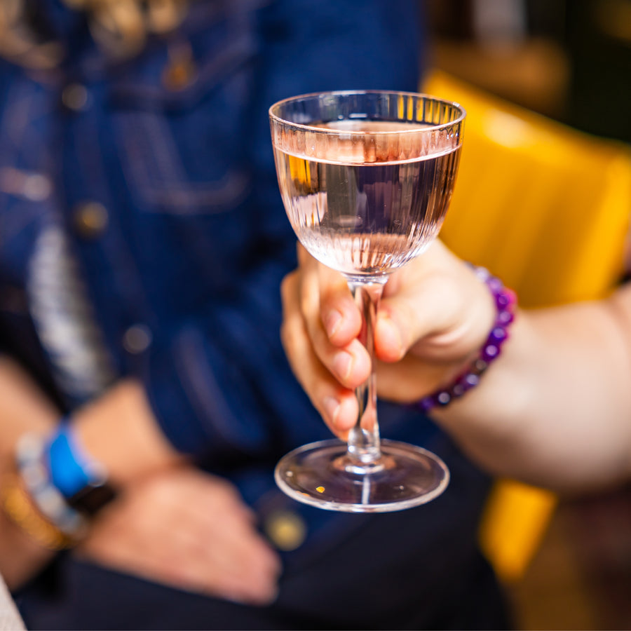 Someone is holding a lychee martini. It looks like they are in a bar. There is someone blurred in the background wearing a blue top and jeans, and sitting on a bright yellow chair.