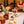 Load image into Gallery viewer, The Aperol Negroni is shown here in a glass being drunk by someone out of shot. There is a Lychee Martini in a nice nic and Nora glass on the table in what could be a house or a bar. There are other drinks and a vase of dried flowers blurred out on the table, and what looks like a bowl of almonds. 
