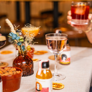 The Aperol Negroni is shown here in a glass being drunk by someone out of shot. There is a Lychee Martini in a nice nic and Nora glass on the table in what could be a house or a bar. There are other drinks and a vase of dried flowers blurred out on the table, and what looks like a bowl of almonds. 