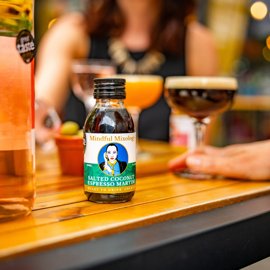 A mini Espresso Martini bottle is on a table in shot, with some blurred cocktails and a person in the background. It's a wooden table, and looks like it is outside.