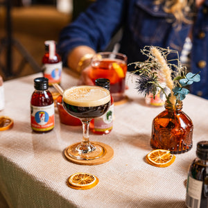 There are some cocktails on a table, with a tablecloth and a vase of dried flowers. It's an espresso martini. A lady is in the background drinking an aperol negroni, but is out of shot. Lots of Mindful Mixology mini cocktails are on the table, with some dried orange slices.