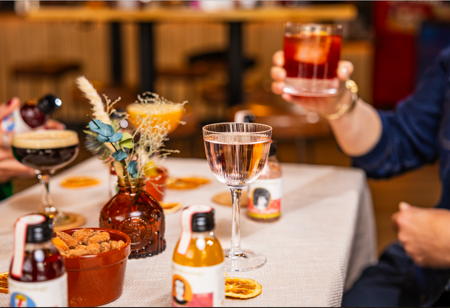 A table with a cream cloth is in a bar with nice lighting. There are lots of cocktails on the table, and mini cocktail bottles, with a bowl of almonds and a vase of dried flowers. There is someone toasting in the background, holding an old fashioned. 